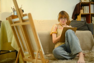 Yaratıcı genç sanatçı yerde oturuyor ve tuvale resim yapıyor..