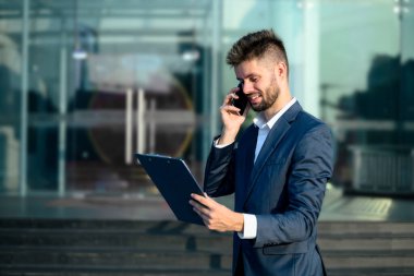 İş adamı elinde bir pano tutuyor ve modern ofis binasının dışında akıllı telefondan konuşuyor..
