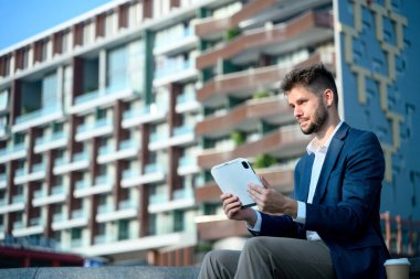 Genç işadamı, modern bir ofis binasının önünde otururken dijital tabletini inceliyor..