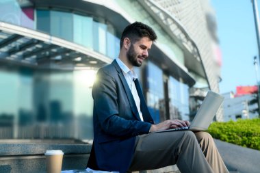 Gülümseyen beyaz iş adamı çağdaş bir şehir manzarasında dizüstü bilgisayarı üzerinde çalışıyor..