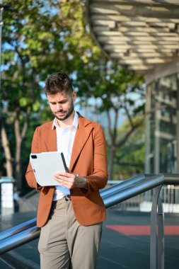 Yakışıklı iş adamı dijital tablet kullanıyor. Çağdaş cam ofis dışındaki parmaklıklara yaslanıyor..