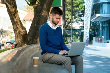 Diz üstü bilgisayarı üzerinde çalışan beyaz erkek serbest çalışan eğimli bir bankta oturuyor..
