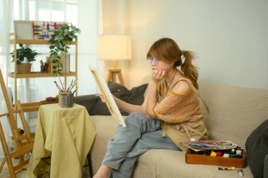 Düşünceli kadın ressam elinde tuval tutarak ev stüdyosunda son eseri üzerinde çalışıyor..