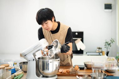 Genç adam kurabiye hamuru hazırlarken tezgah karıştırıcısına malzeme ekliyor..