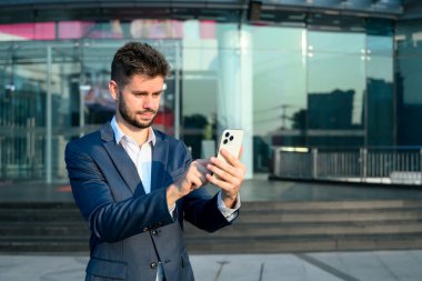 Genç işadamı modern cam ofis binasının önünde duruyor ve akıllı telefonunu kullanıyor..