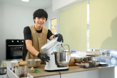 Gülümseyen genç fırıncı kurabiye hamuru hazırlıyor ve mutfakta karıştırıcı kullanıyor..