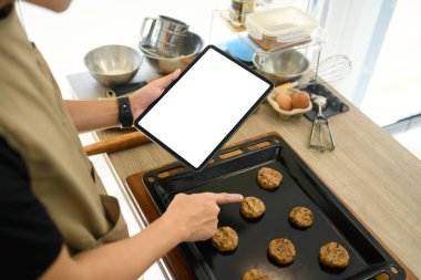 Çikolata parçalı kurabiyeler hazırlarken elinde boş ekranla dijital tablet tutan genç girişimci.