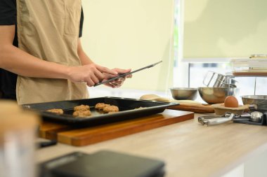 Genç adam mutfakta çikolatalı kurabiye hazırlıyor ve dijital tablette tarifi takip ediyor..