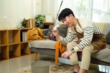 Önlüklü bir adam dağınık bir oturma odasında oturuyor ve buruşuk kağıtları çöpe atıyor..