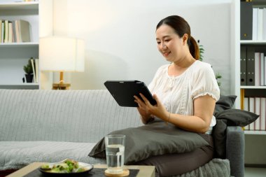 Oturma odasında yemek yerken dijital tablet kullanan gülümseyen Asyalı kadın..