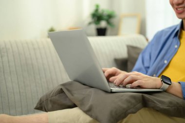 Gülümseyen Asyalı genç adam rahat bir oturma odasında dizüstü bilgisayarda çalışıyor ya da geziniyor..