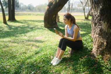 Spor giyimli genç bir kadın parkta ağacın altında oturuyor ve akıllı telefon kullanıyor..