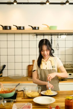 Önlük takan genç bir kadın yeni pişmiş spagettiyi ev mutfağında bir tabağa koyar..