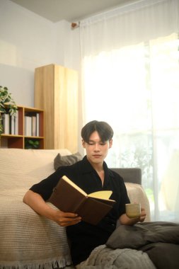 Genç Asyalı adam oturma odasında sıcak bir şeyler içerken kitap okuyor..