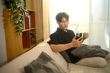 Telsiz kulaklık takan ve kahve içerken akıllı telefon kullanan mutlu genç adam..