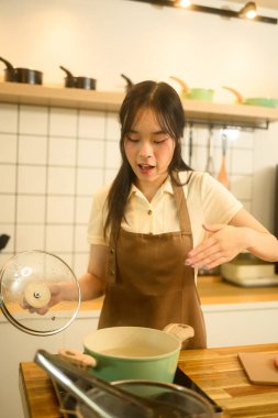 Kahverengi önlüklü genç kadın haşlanmış makarnayı kontrol ederken tencerenin kapağını kaldırıyor..