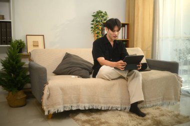 Rahat Asyalı bir adam modern bir oturma odasında rahat bir koltukta oturuyor ve dijital tablet kullanıyor..