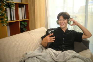 Kablosuz kulaklık takan neşeli Asyalı genç adam akıllı telefondan bir şeyler izliyor..