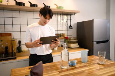 Genç Asyalı bir adam protein içeceği hazırlıyor ve tahta tezgahta dijital tablet kullanıyor..