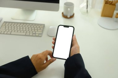 İş kadınının elinde beyaz ekranlı akıllı telefon tutarak iş yerinde oturmasına yakın çekim.