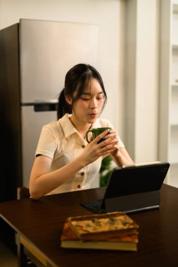 Mutfaktaki ahşap masada oturan, dijital tablet üzerinde kahve yudumlayan genç bir kadın..