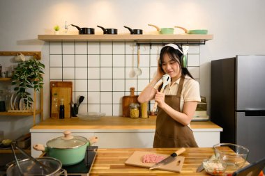 Kulaklık takan neşeli kadın mutfağında yemek pişirmekten hoşlanır..