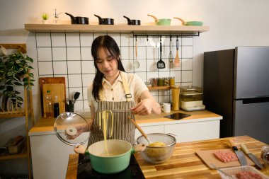 Önlüklü genç bir kadın tencerede taze haşlanmış spagetti yemek için maşa kullanıyor. Ev yemeği konsepti.
