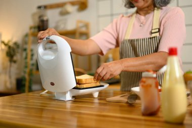 Önlük giymiş yaşlı bir kadın sıcak mutfakta kahvaltı hazırlıyor..