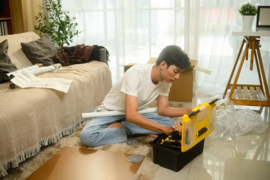 Genç Asyalı adam oturma odasında yeni mobilya monte ederken alet arıyor..