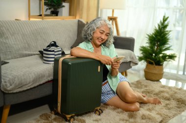 Neşeli olgun kadın bavulunun yanında yerde oturuyor ve akıllı telefon kullanıyor..