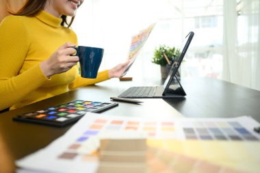 Yaratıcı tasarımcı elinde kahve fincanı tutuyor. Masadaki bir proje için renk seçimi üzerinde çalışıyor..