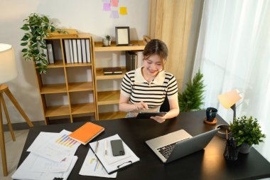 Masasında dijital tablet kullanan neşeli genç bir kadın. Etrafı mali raporlar ve dizüstü bilgisayarla çevrili..