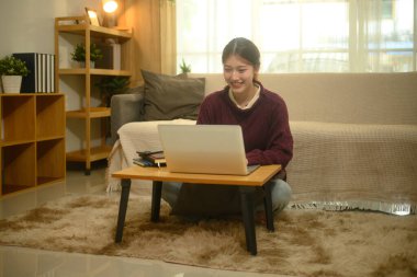 Gülümseyen Asyalı kadın, dizüstü bilgisayarıyla çalışıyor, oturma odasında halıda oturuyor..
