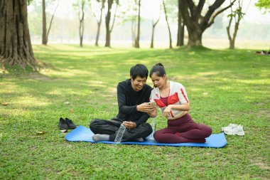 Yeşil bir parkta oturmuş spor salonlarından sonra cep telefonlarını kontrol eden mutlu bir çift..