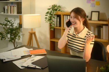 Neşeli genç girişimci iş yerinde oturmuş telefonla konuşuyor..