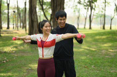 Erkek, güneşli bir parkta dambıl egzersizinde kadına yardım ediyor..