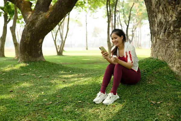 Ağacın altında oturan, elinde kablosuz kulaklıkla akıllı telefon kullanan genç bir kadın..