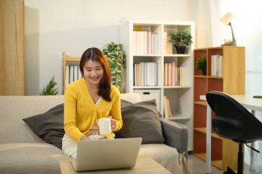 Gülümseyen genç bir kadın elinde bir fincan sıcak çay ile oturma odasında dizüstü bilgisayarla çalışıyor..