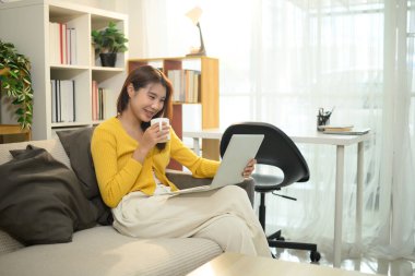 Neşeli Asyalı kadın elinde kahve fincanıyla oturma odasında laptopunu karıştırıyor..