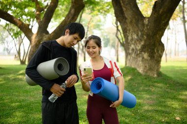 Yoga minderleri sarılı mutlu çift parkta dikiliyor ve akıllı telefondaki fitness uygulamasını kontrol ediyor..