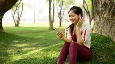 Parktaki bir ağacın altında oturmuş gülümseyen genç bir kadın. Telefonunu ve kulaklıklarını kullanıyor..