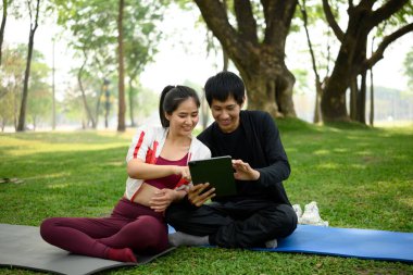 Asyalı çift dijital tablet üzerinde egzersiz planlarını kontrol ediyor, güneşli bir parkta yoga minderlerinin üzerinde oturuyorlar..