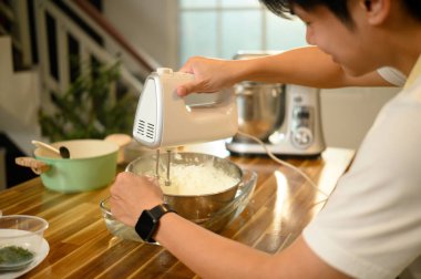 Önlüklü genç adam, kremayla karıştırıcıyı paslanmaz çelik bir kasede karıştırıyor..