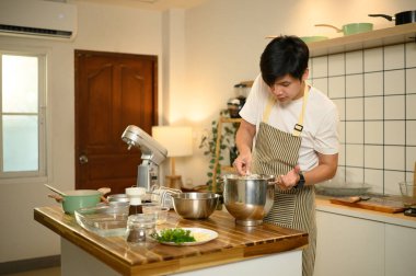 Genç Asyalı adam, parlak bir mutfakta çırpıcıyla paslanmaz çelik kasede krema hazırlıyor..