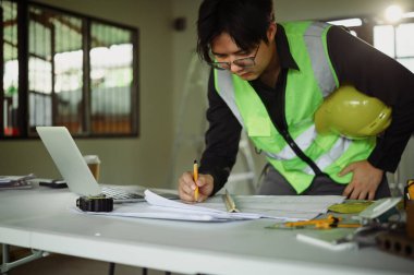 Güvenlik yeleği giymiş bir mühendis bir masada inşaat planları üzerinde çalışıyor..