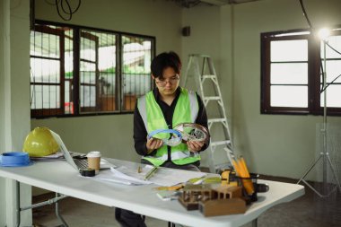 Yansıtıcı güvenlik yeleği giymiş bir mühendis inşaat alanındaki elektrik kablolarını inceliyor..