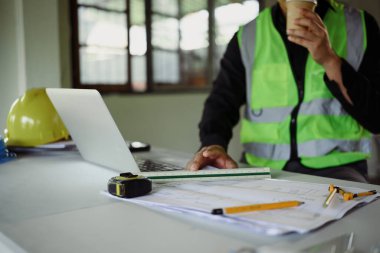 Bir dizüstü bilgisayardaki inşaat planlarını incelerken şantiyede kahve molası veren bir mühendis..