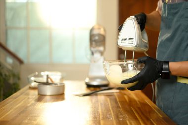 Siyah eldivenli adam cam kasede kremayla elektrikli el mikserini karıştırıyor..