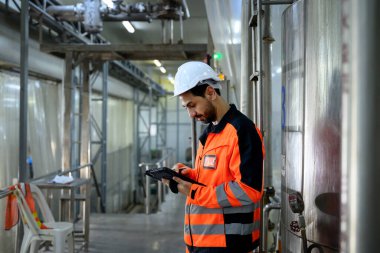 Üretim tesisinde endüstriyel ekipmanları denetlemek için dijital tablet kullanan koruyucu üniformalı bir fabrika mühendisi..