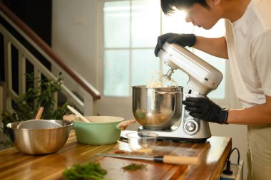 Genç Asyalı adam modern bir mutfakta çırpılmış krema yapmak için karıştırıcı kullanıyor..